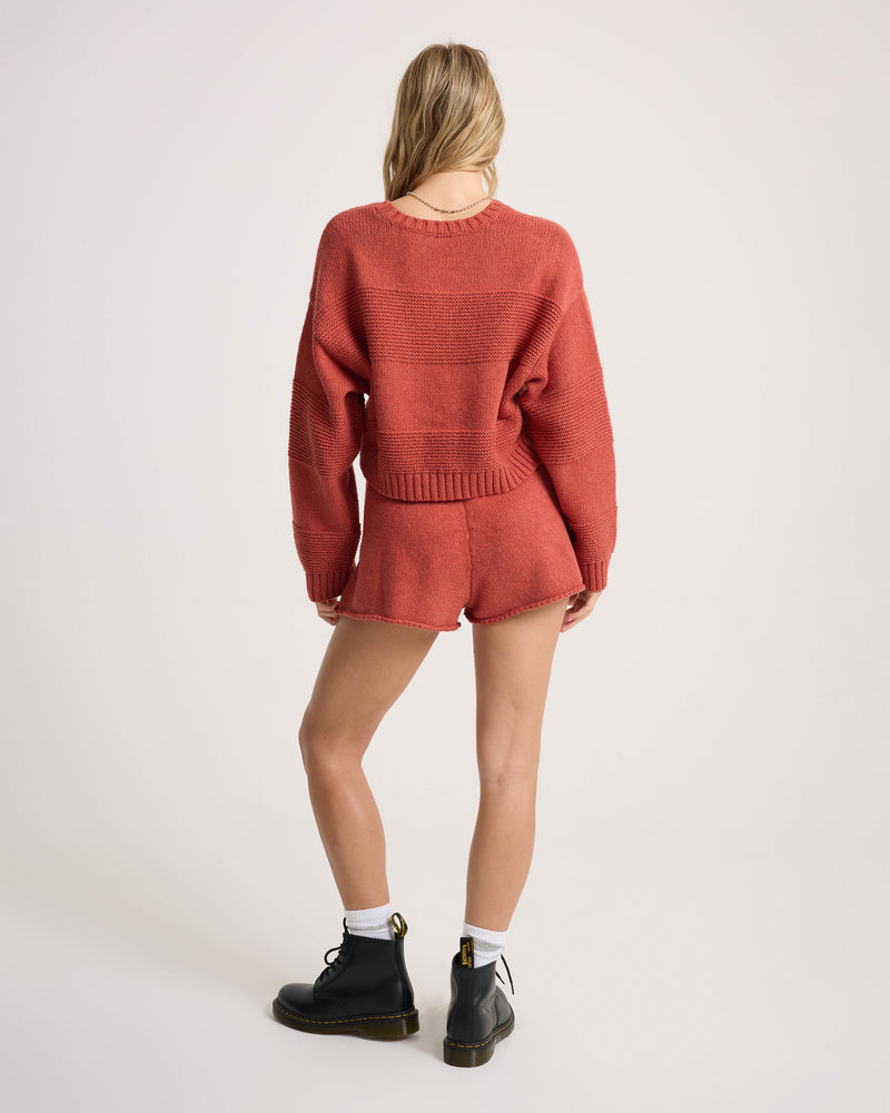 A woman stands facing away, wearing the Volcom Women’s Dayz Like This Sweater—a matching rust-colored cropped crew sweater and shorts set—paired with black ankle boots and white socks. The background is plain and light-colored.