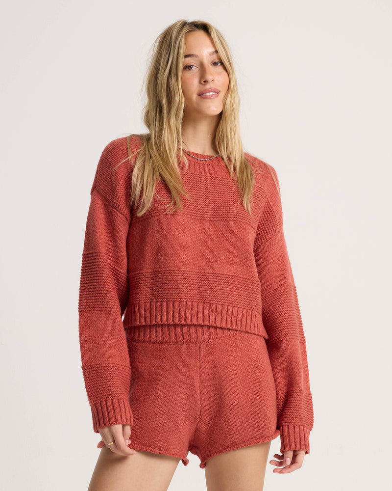 A woman with long blonde hair wears the Volcom Women’s Dayz Like This Sweater and matching shorts, featuring variegated red stripes, as she stands against a plain light background and smiles softly at the camera.