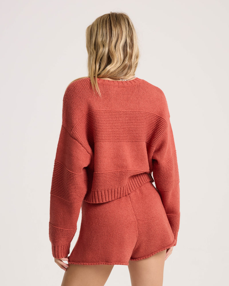 A person with wavy blonde hair, seen from behind, is wearing the Volcom Women’s Dayz Like This Sweater—a loose, cropped crew knit with variegated stripes—paired with matching shorts against a plain light background.