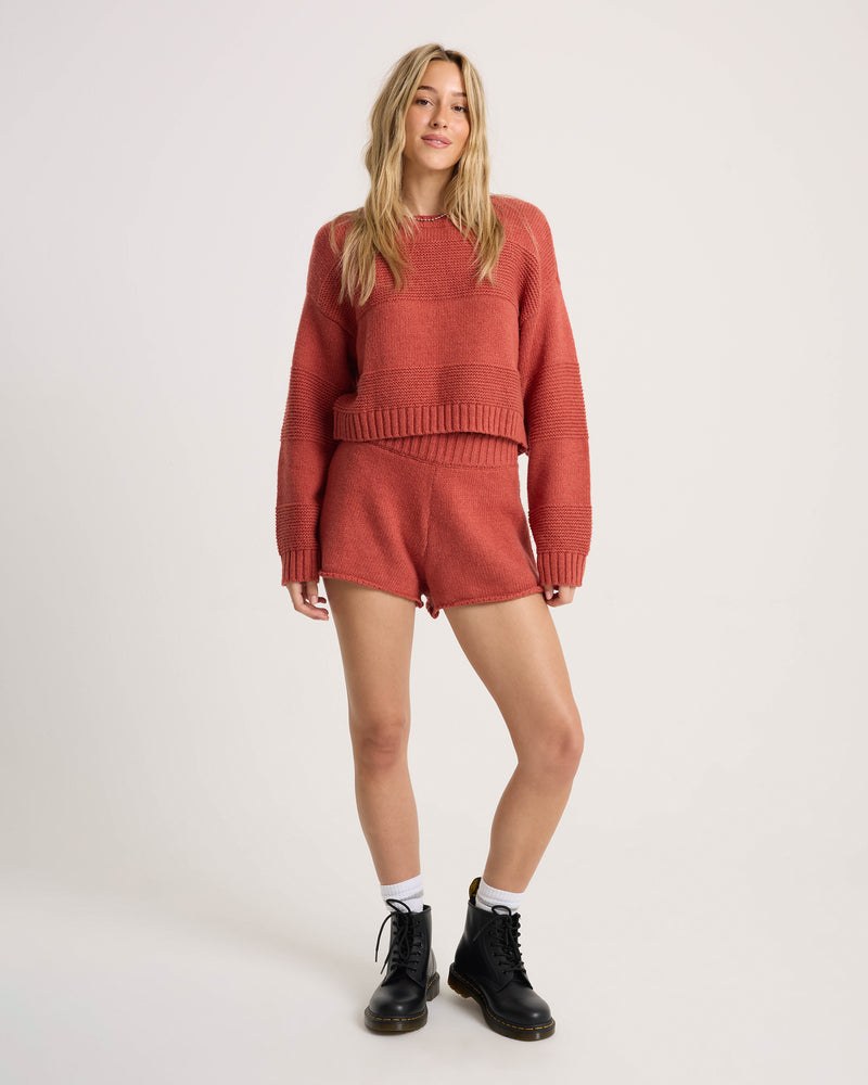 A woman stands against a plain background, wearing the Volcom Women’s Dayz Like This Sweater—a red variegated stripe knit with dolman sleeves—paired with matching shorts, white socks, and black boots. The cropped crew sweater perfectly complements her long blonde hair and relaxed expression.