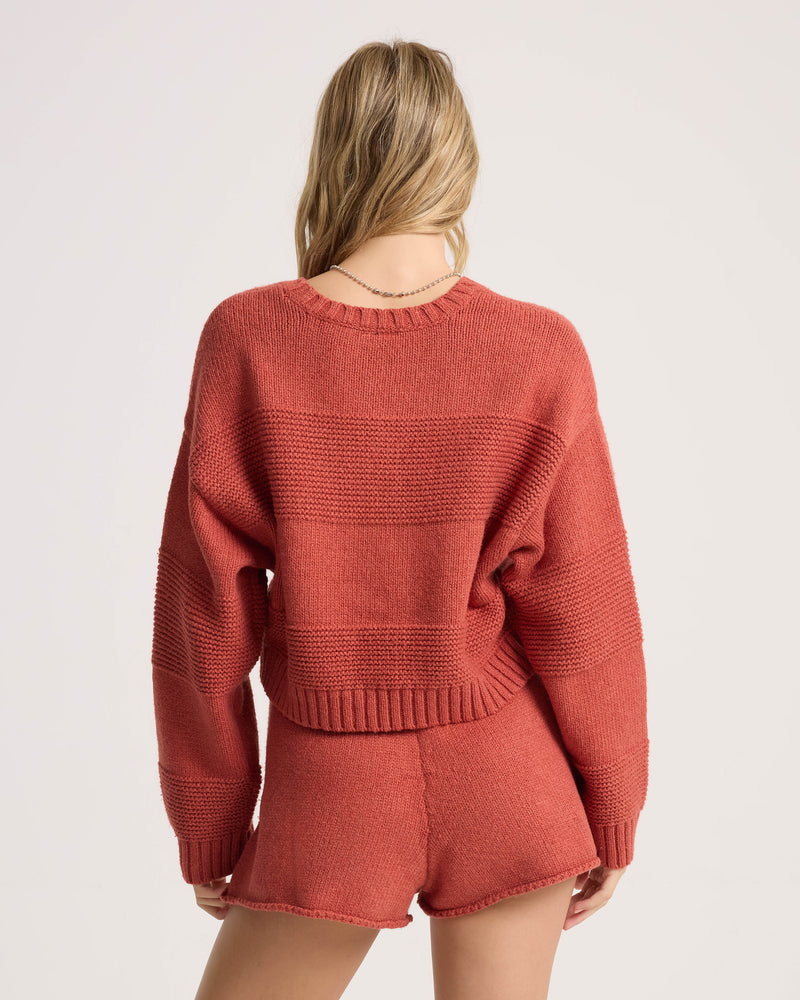 A woman with long blonde hair is wearing the Volcom Women’s Dayz Like This Sweater—a rust-colored, cropped crew style with dolman sleeves—paired with matching shorts. She stands with her back to the camera against a plain light background.