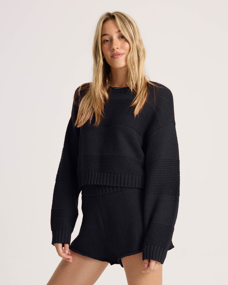 A woman with long blonde hair wears the Volcom Women’s Dayz Like This Sweater in black, paired with matching black shorts, standing against a plain light-colored background.