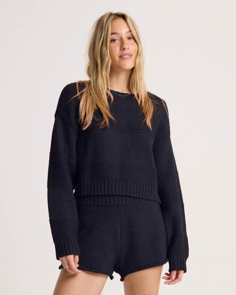 A woman with long blonde hair wears the Volcom Women’s Dayz Like This Sweater in black, paired with matching black knit shorts, standing against a plain light background.