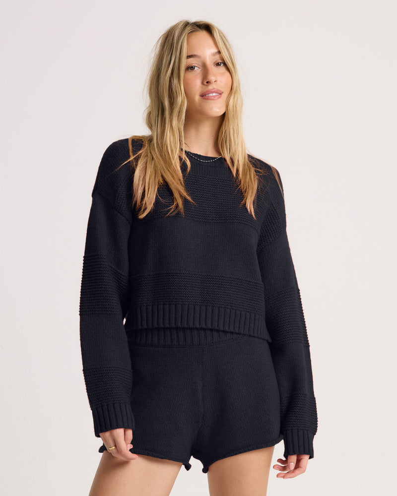 A woman with long blonde hair wears the Volcom Women’s Dayz Like This Sweater in black, paired with matching black knit shorts, standing against a plain light background.