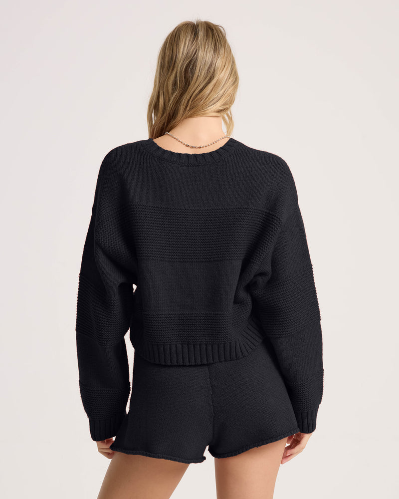 A woman with long blonde hair is standing with her back to the camera, wearing the Volcom Women’s Dayz Like This Sweater in black, paired with matching black shorts against a plain light background.