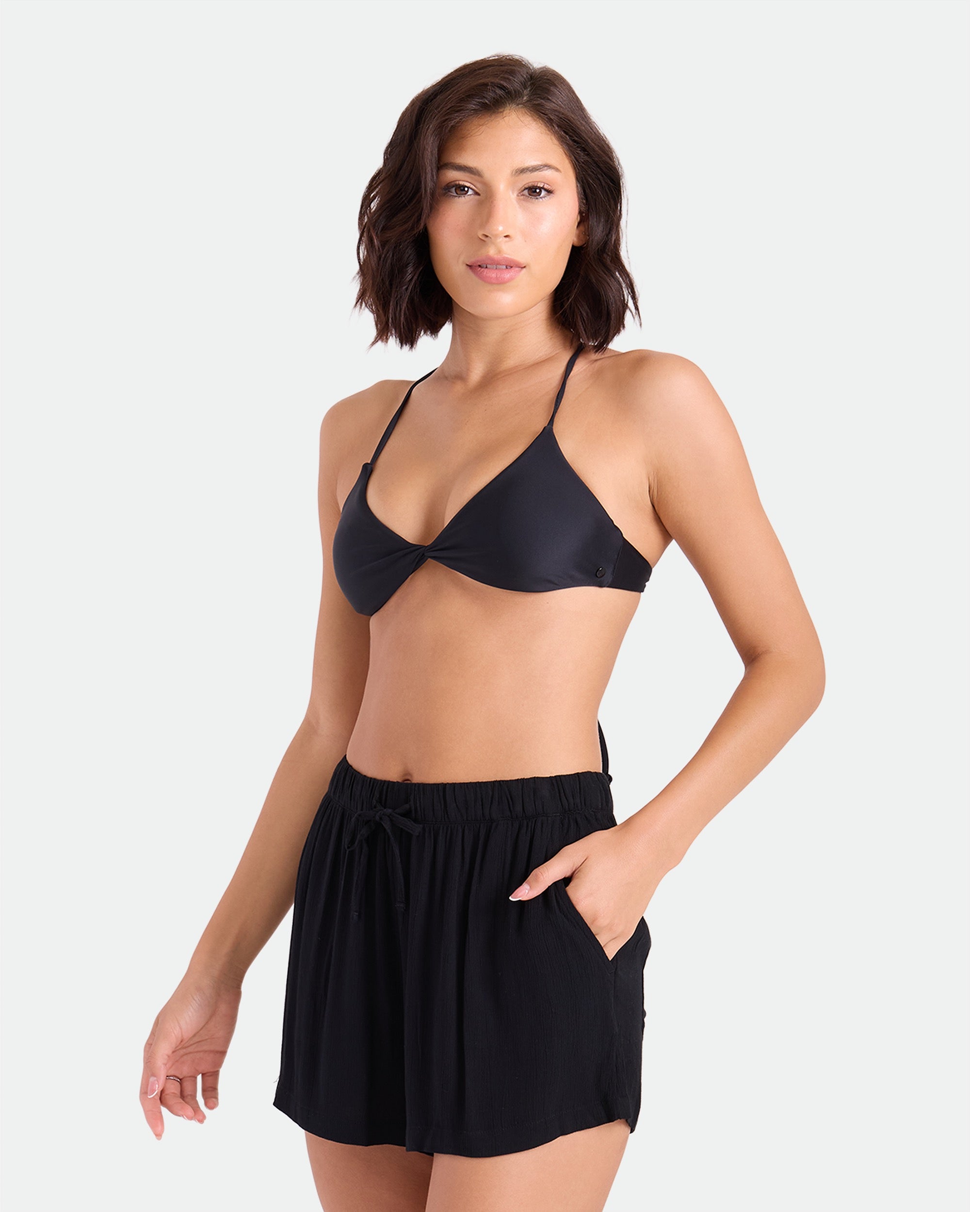 A woman with short brown hair wears a black bikini top and Volcom Women's Stone Shine Shorts Vsc with an elastic waistband, standing against a plain light background with one hand in her pocket.