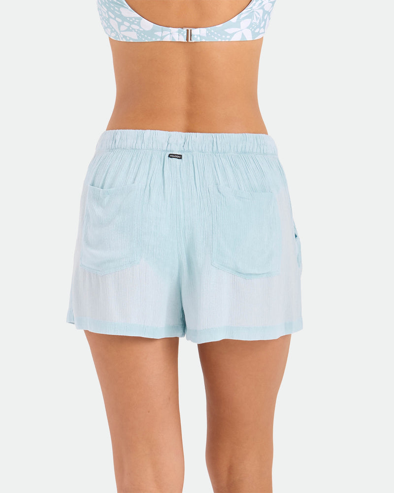A woman is seen from behind wearing the Volcom Women's Stone Shine Shorts Vsc in light blue, made of breathable viscose with an elastic waistband and two back pockets, paired with a patterned bikini top.