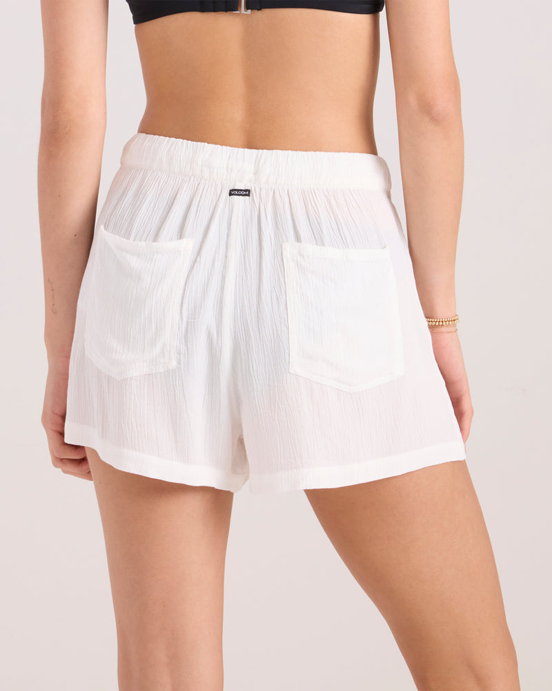 A woman is seen from the back wearing a black bikini top and Volcom Women's Stone Shine Short Vsc in Star White. The lightweight viscose shorts have a crinkled texture, elastic waistband, and two back pockets, showing her lower back and legs.