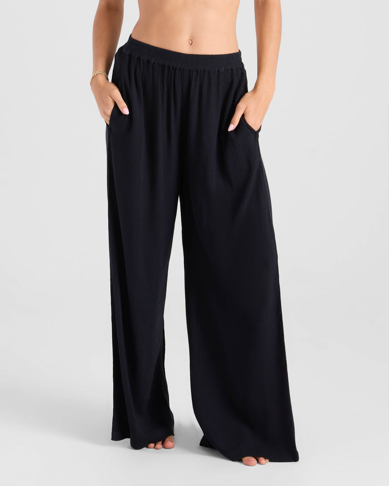 A barefoot person models the Volcom Women's Stoneshine Junki Pants—loose-fit, high-waisted black wide leg with elastic waist and pockets. The draped viscose fabric flows elegantly. Upper body cropped; plain light gray background.