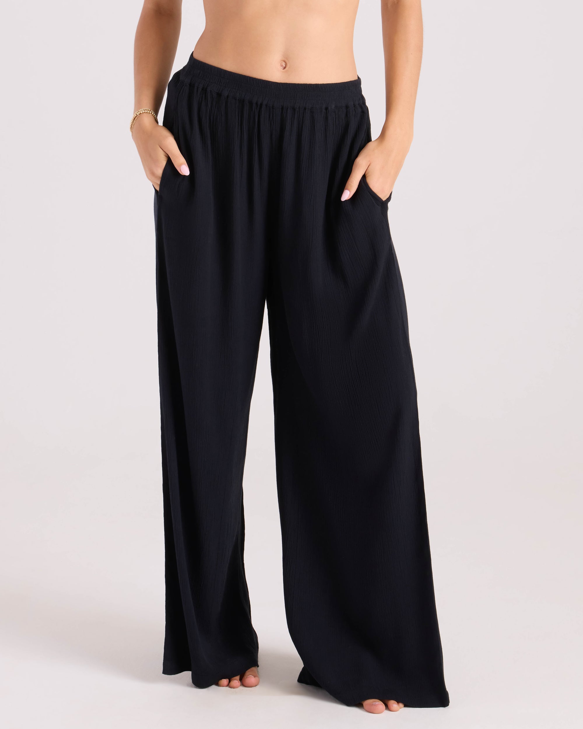 A person stands barefoot against a plain light background, wearing Volcom Women’s Stoneshine Junki Pant—high-waisted, loose-fitting black wide leg pants with an elastic waistband and hands in the pockets.