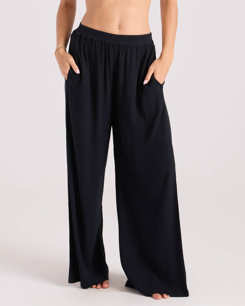 A person stands barefoot against a plain light background, wearing Volcom Women’s Stoneshine Junki Pant—high-waisted, loose-fitting black wide leg pants with an elastic waistband and hands in the pockets.