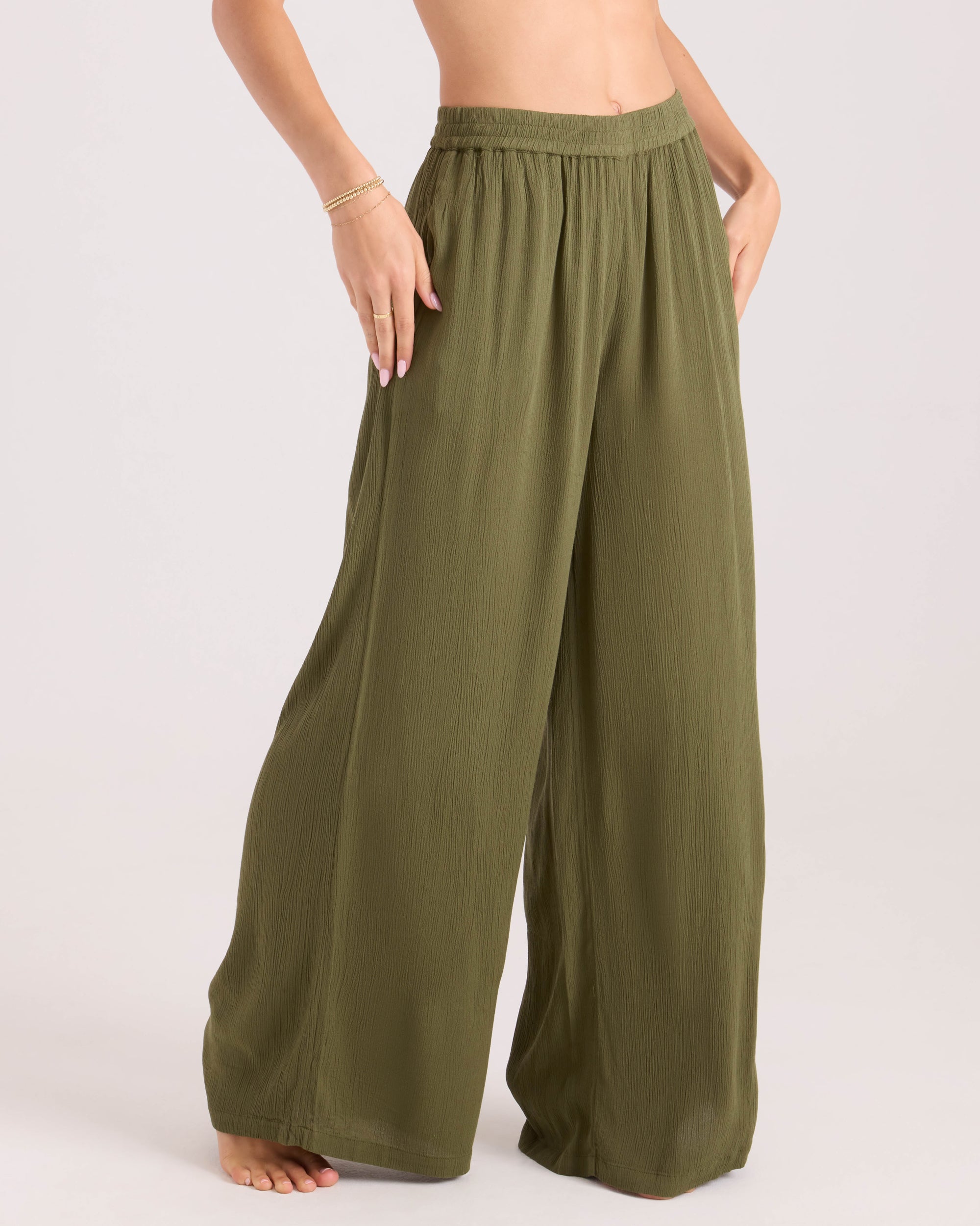 A person stands barefoot against a plain light background, wearing Volcom Women's Stoneshine Junki Pant in olive green. The wide-leg pants feature a flowy, textured fabric, elastic waistband, and hands resting at the waist and hip.
