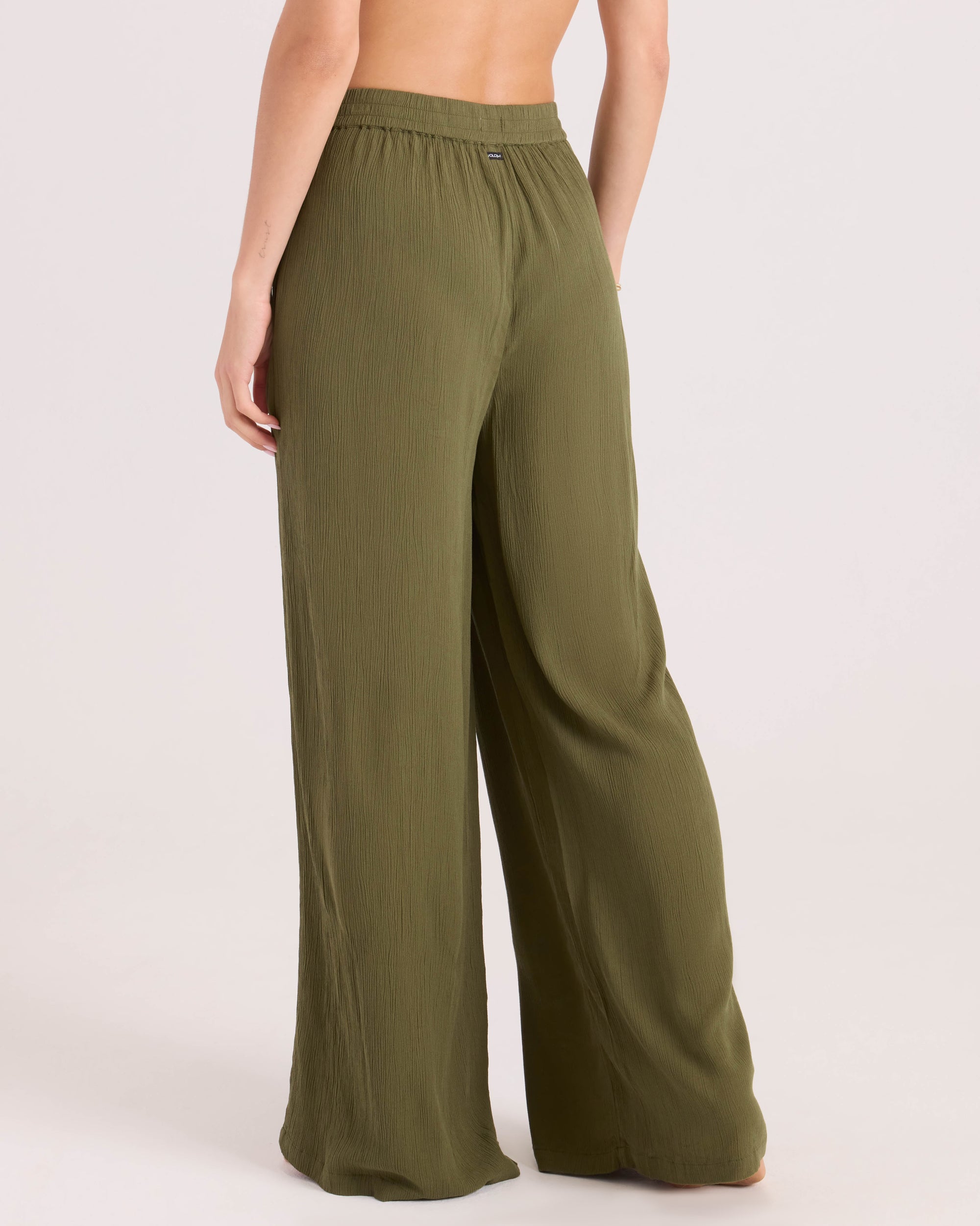 A person faces away, wearing Volcom Women's Stoneshine Junki Pant—loose, high-waisted olive green pants with a crinkled texture, relaxed fit, and wide legs. The upper body is not fully visible.