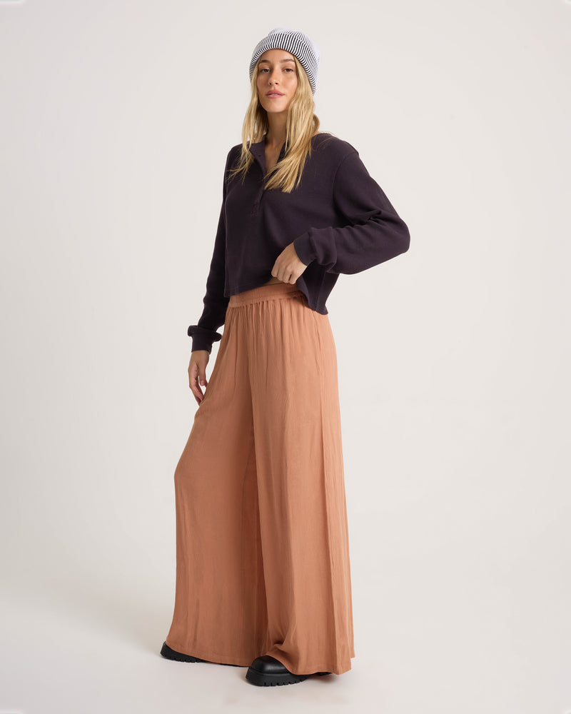 A person wears a dark long-sleeve polo shirt, a striped beanie, and Volcom Women’s Stoneshine Junki Pant in a relaxed-fit, wide-leg peach style with black boots.