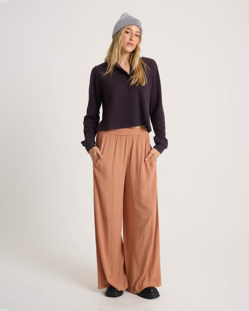 A person wears relaxed fit, high-waisted, wide-leg peach Volcom Women’s Stoneshine Junki Pants, a dark long-sleeve henley shirt, and a striped knit beanie.