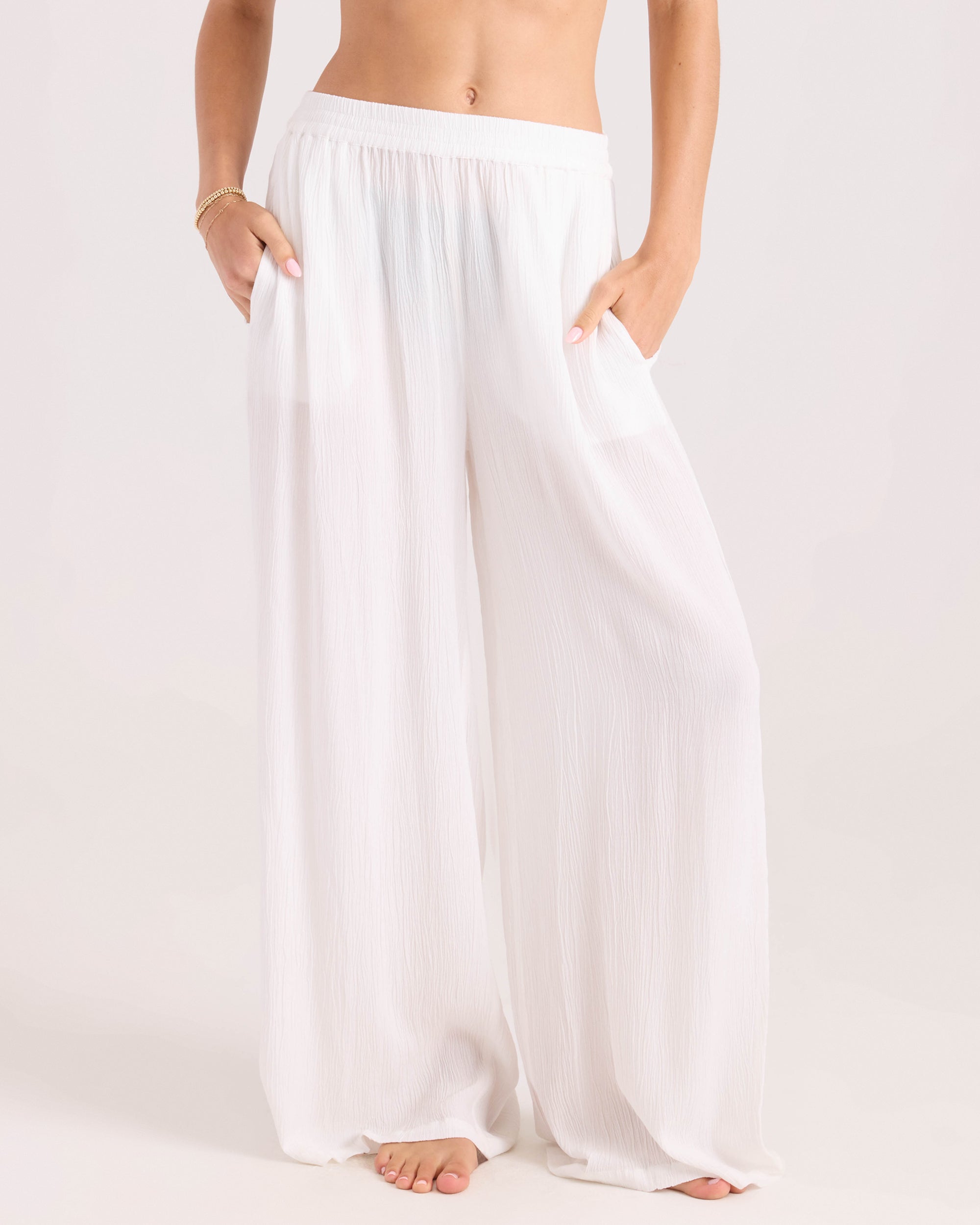 A person stands barefoot, hands in pockets, wearing Volcom Women's Stoneshine Junki Pant—loose, high-waisted, wide-leg white pants in lightweight fabric. Image is cropped at the waist and ankles.