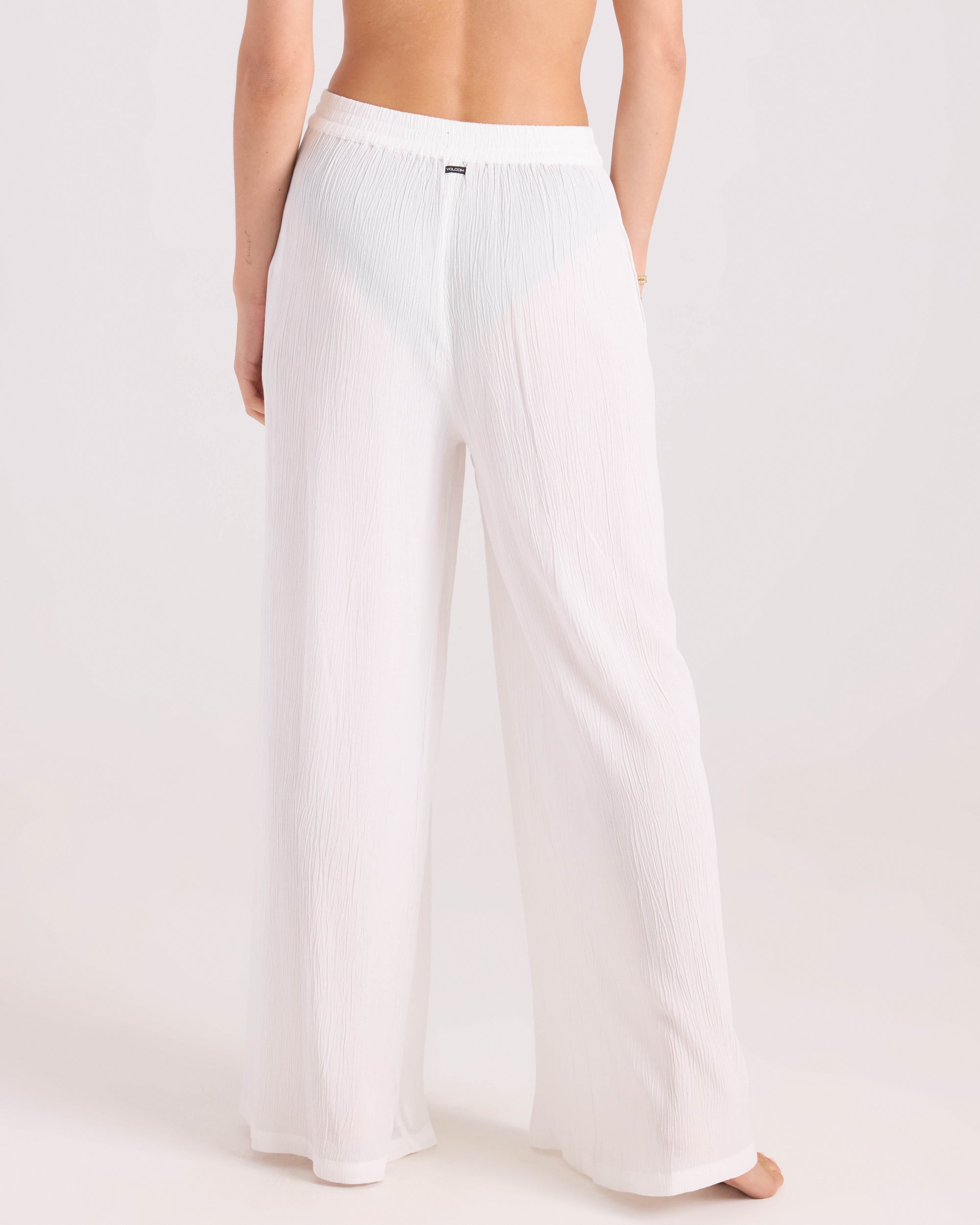 A person is shown from the back wearing Volcom Women's Stoneshine Junki Pant—loose, white, semi-sheer wide-leg pants with an elastic waistband. The plain, light background partially shows their upper body and feet.