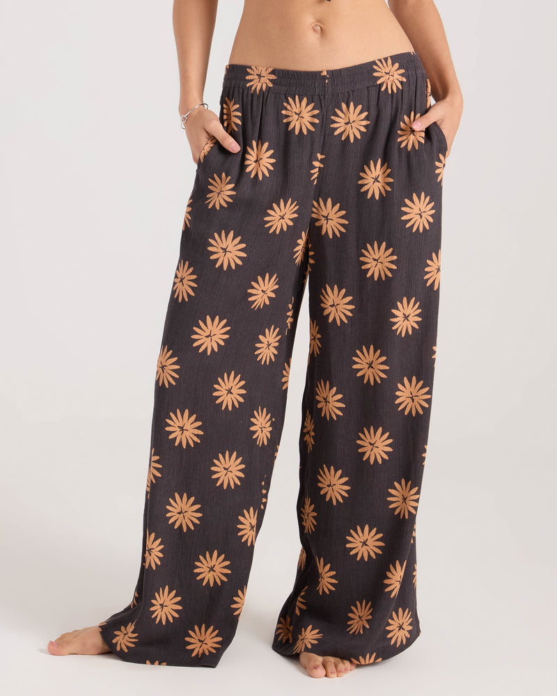 A person stands barefoot wearing the Volcom Women's Midnite Ramble Pant, which features a high-waisted, wide leg design with an all-over orange floral pattern. Their hands are in the pockets and their upper body is mostly out of frame.