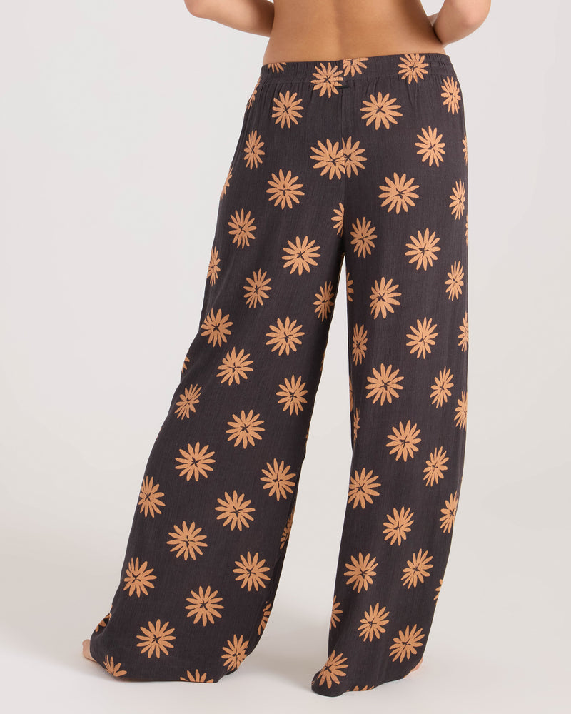 A person stands barefoot, facing away from the camera against a plain white background, wearing Volcom's Women's Midnite Ramble Pant—high-waisted, wide leg pants with an orange floral pattern.