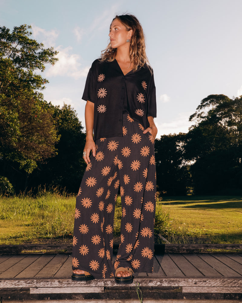 A person with wavy hair stands outdoors wearing a loose black shirt and Volcom Women's Midnite Ramble Pants, which are high-waisted with a tan starburst pattern, paired with black sandals amid green trees under a partly cloudy sky.