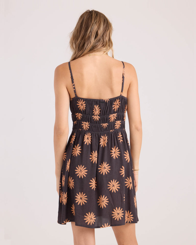 A woman with wavy blonde hair is seen from behind wearing the Volcom Women's Midnite Ramble Dress, a sleeveless black mini dress with thin straps, a gathered waist, and an orange daisy floral pattern.