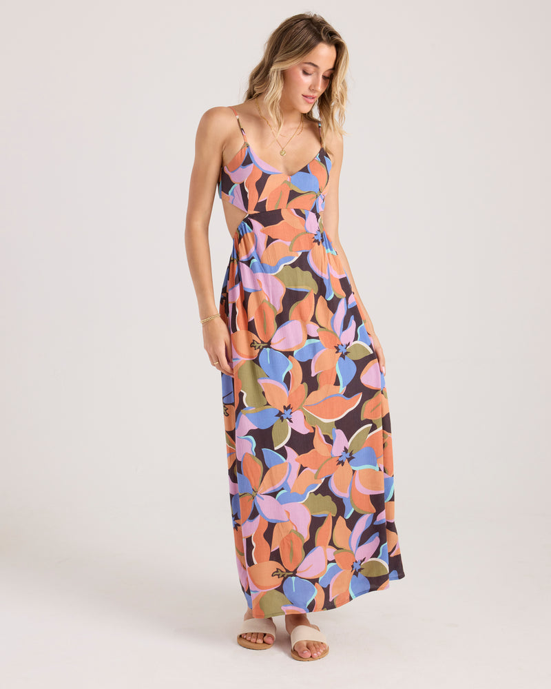 A woman wearing the Volcom Women's Post Punk Dress stands against a plain light background. The dress features an abstract leaf print and A-line maxi silhouette with thin straps. She has light brown hair, sandals, and touches her hair while looking down.