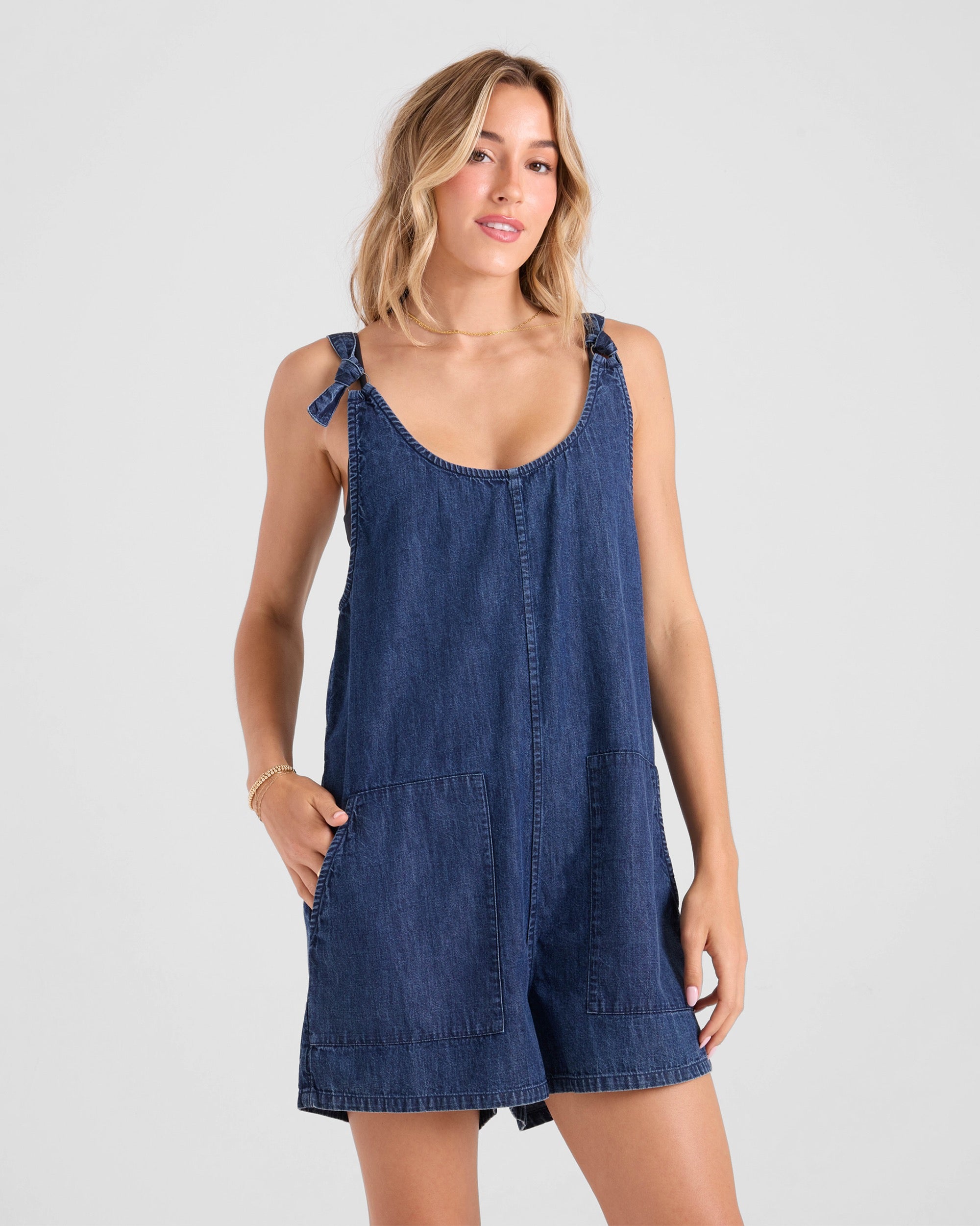 A woman with light wavy hair poses against a plain background wearing the Volcom Women's Lived In Shorts Romper, a utility-style sleeveless denim outfit with tie straps and front pockets. She stands relaxed and smiles subtly.