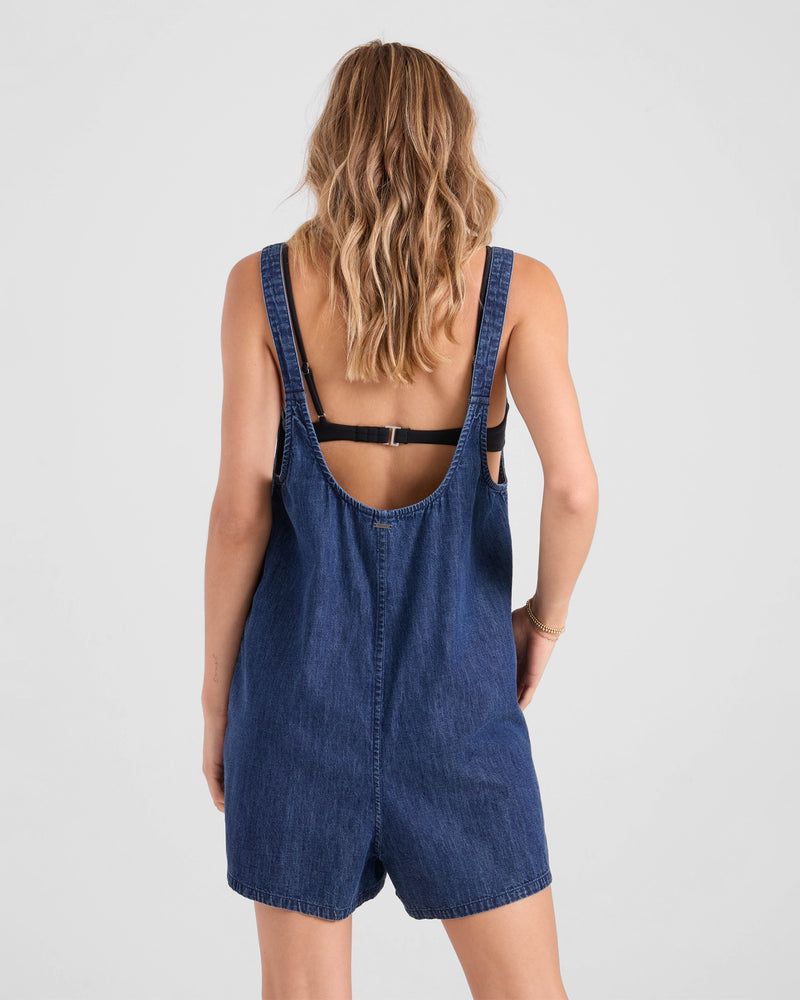 A woman with wavy, shoulder-length hair stands with her back to the camera, wearing the Volcom Women's Lived In Shorts Romper—crafted from 100% cotton denim—over a black bra against a plain, light background.