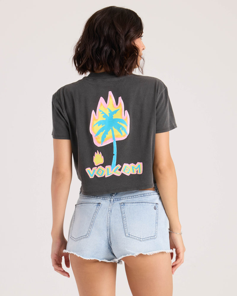 A woman with wavy brown hair wears the Volcom Women's Pocket Dial Tee—a vintage black cropped crew neck with a colorful flaming palm tree and VOLCOM text on the back—paired with light denim shorts, shown from behind against a plain background.