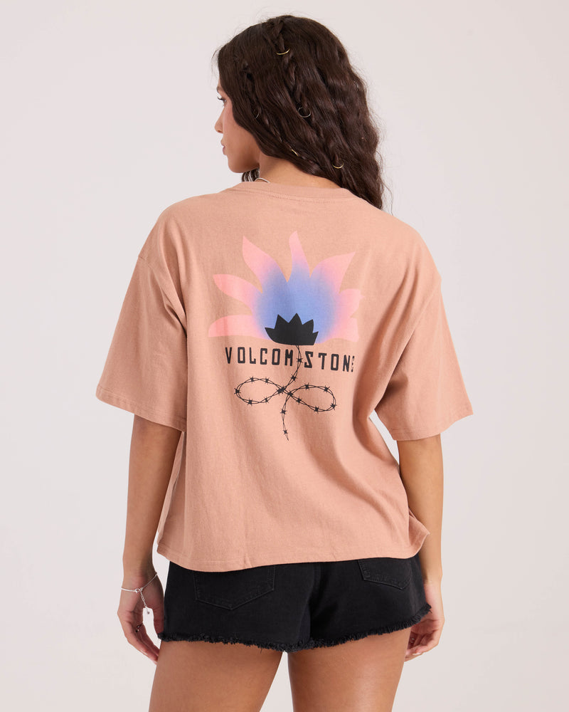 A person with long wavy hair, facing away, wears the Volcom Women's Bliss Tee in peach with a graphic pink and blue flower, black crown, and VOLCOM STONE text on the back, paired with black denim shorts.