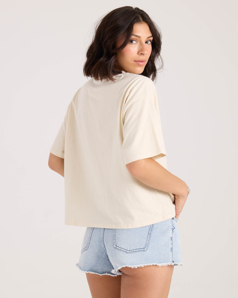 A woman with shoulder-length dark hair models the Volcom Women's Bliss Tee and light blue denim shorts. She stands with her back to the camera, glancing over her shoulder against a plain light background.