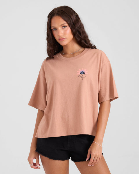 A young woman with wavy brown hair wears the Volcom Women's Burnout BF Tee, a loose light brown T-shirt with a small flower graphic on the chest, paired with black shorts. She stands against a plain light gray background, gazing calmly at the camera.