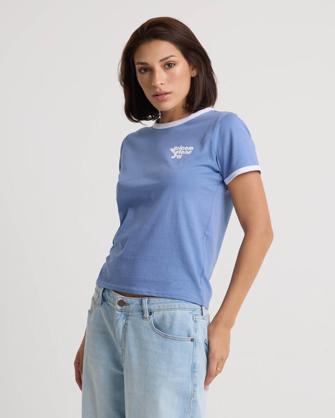 A woman with straight brown hair wears the Volcom Women's Brk Up Ringer Tee, a Horizon Blue crew neck t-shirt with white trim, paired with faded blue jeans against a plain white background.