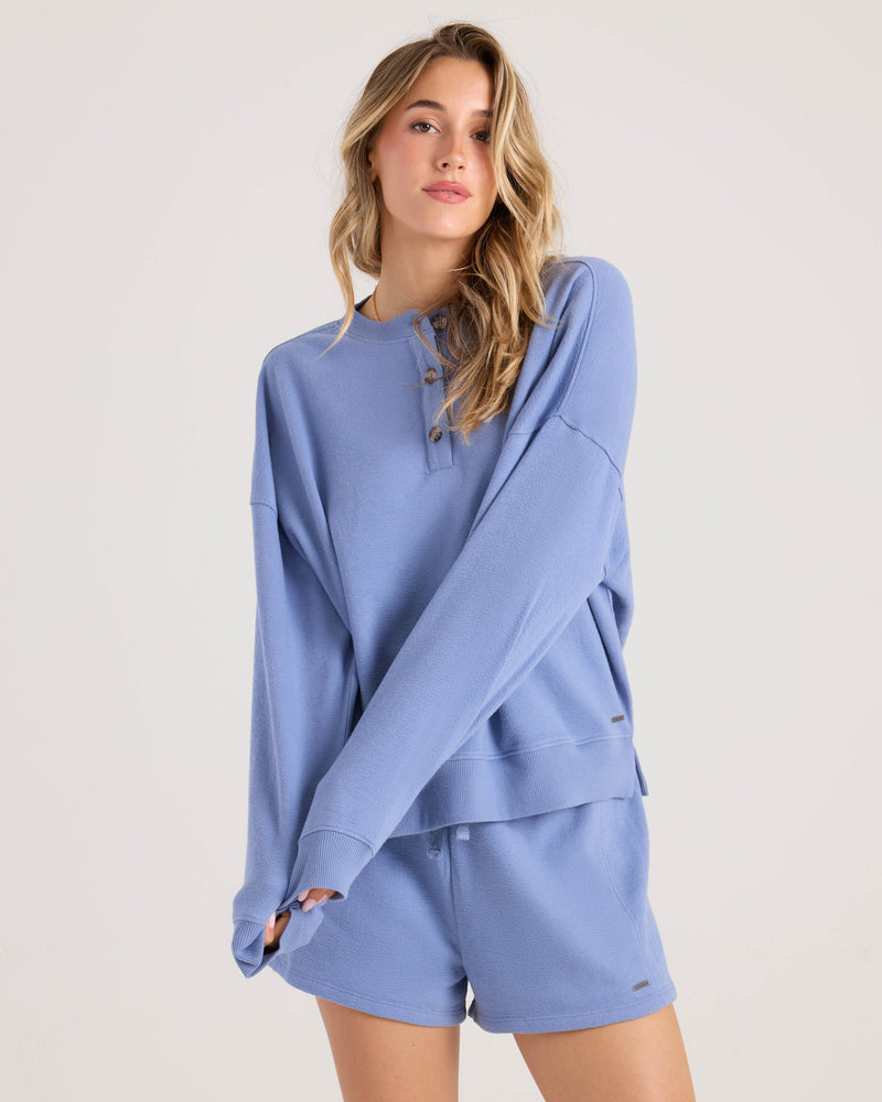 A woman with long, wavy blonde hair models the Volcom Women's Lived In Fleece Henley in a relaxed fit, light blue with buttons, paired with matching shorts against a plain light background.