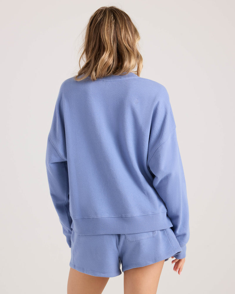 A woman with wavy, shoulder-length hair is seen from the back wearing the Volcom Women's Lived In Fleece Henley in light blue and matching shorts, standing against a plain light background.