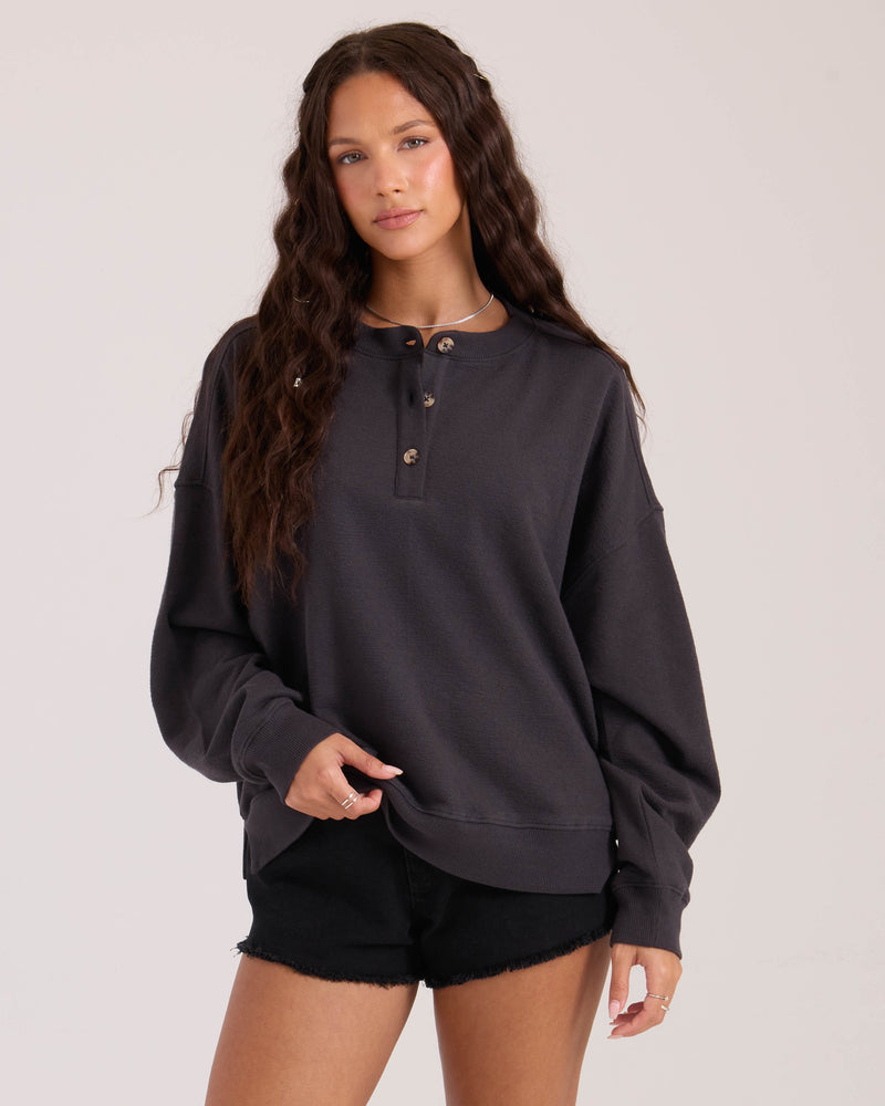 A woman with long wavy brown hair wears the Volcom Women's Lived In Fleece Henley and black shorts. She stands against a plain light background, looking at the camera with a relaxed expression.