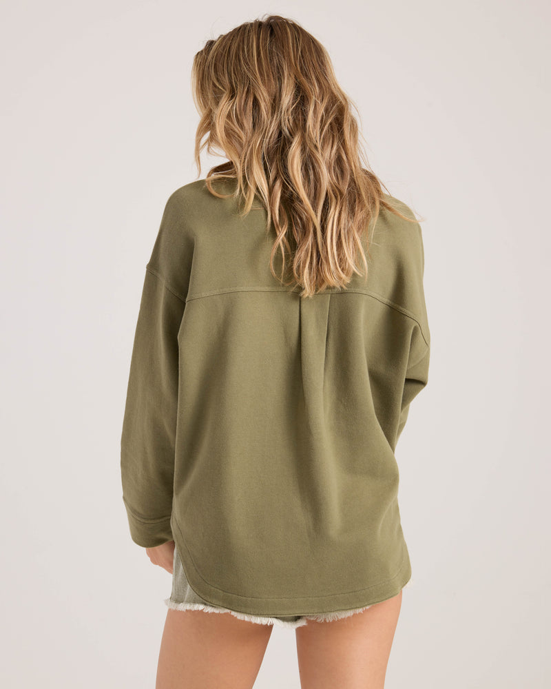 A woman with wavy, light brown hair stands with her back to the camera, wearing the Volcom Women's Nollie Jacket in olive green and light-colored shorts against a plain, light background.