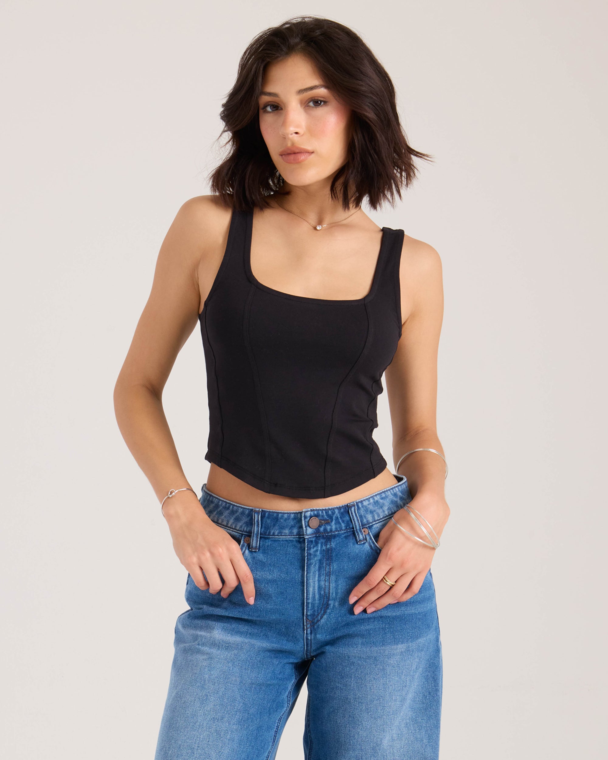 A woman with shoulder-length dark hair wears the Volcom Women's Hot Girl fitted tank and blue jeans, standing against a plain light background. She has a neutral expression and minimal jewelry on her wrists and fingers.