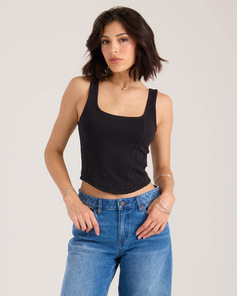 A woman with shoulder-length dark hair wears the Volcom Women's Hot Girl fitted tank and blue jeans, standing against a plain light background. She has a neutral expression and minimal jewelry on her wrists and fingers.