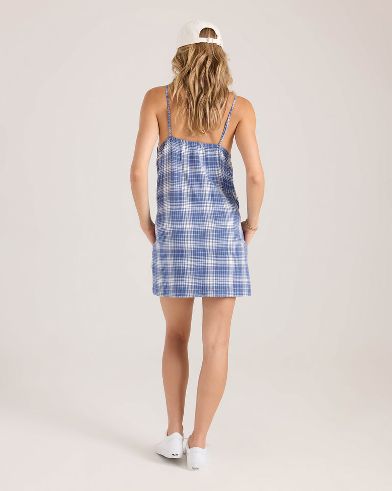 A woman with wavy blonde hair, wearing a white cap and white shoes, models the Volcom Women's Folkin Stone Dress—a blue plaid A-line mini with adjustable straps—while standing facing away in a light studio setting.