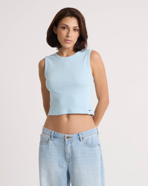 A person with short brown hair wears the Volcom Women's Mary Mack Tank in light blue and matching stretchy knit jeans. They stand against a plain white background, facing the camera with a neutral expression and hands behind their back.