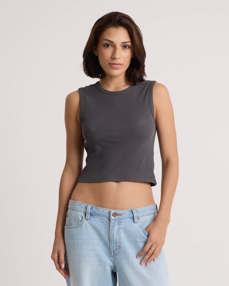 A woman with shoulder-length brown hair wears the Volcom Women's Mary Mack Tank in Vintage Black, featuring a fitted crew-neck design crafted from a soft cotton blend, paired with light blue jeans. She stands against a plain light background with a neutral expression.