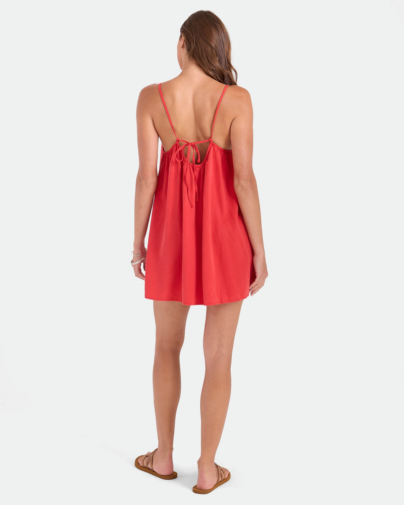 A woman faces away in Volcom's Women's Baby Cakes Mini, a vibrant red dress with thin straps, open tie-back, and breezy A-line shape. She pairs it with brown flat sandals against a plain white background.