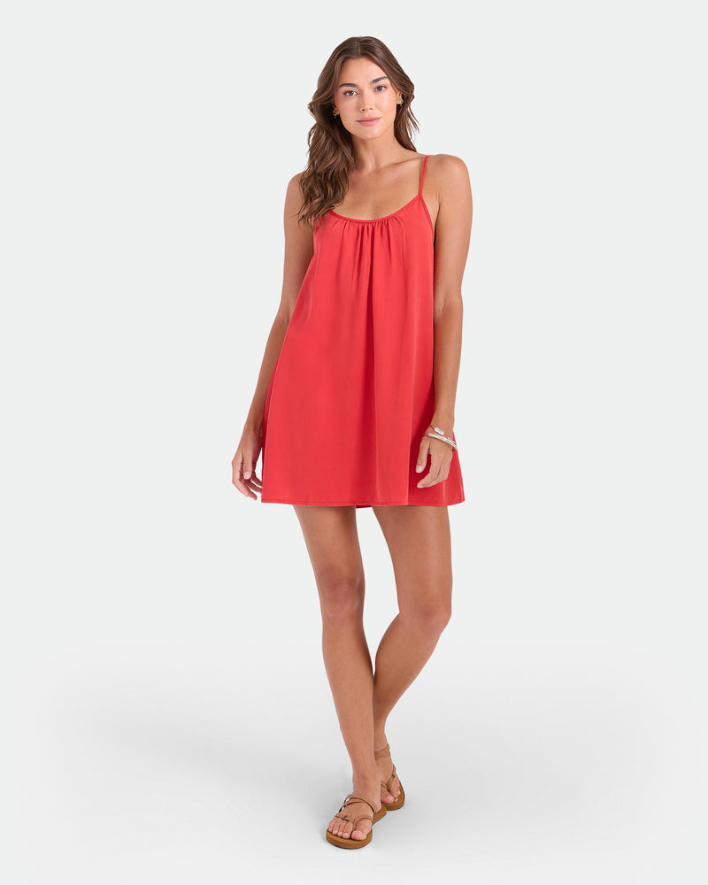 A woman in the Volcom Women's Baby Cakes Mini dress and tan sandals stands against a white background, her long wavy brown hair framing her face as she poses with one hand on her hip and a relaxed expression.