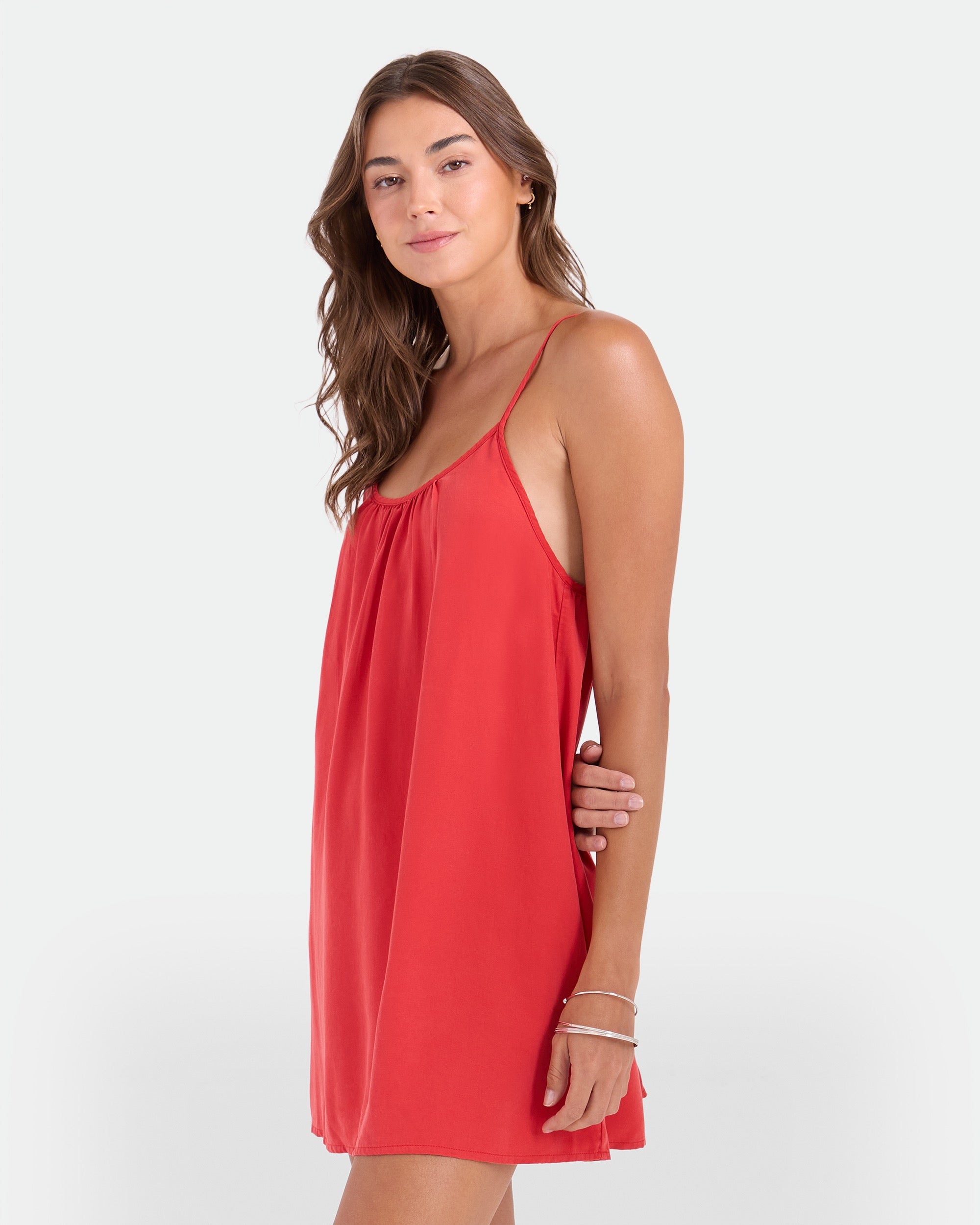 A woman with long brown hair wears the Volcom Women's Baby Cakes Mini, a loose, sleeveless red woven dress with thin straps, while standing against a plain white background and smiling softly.
