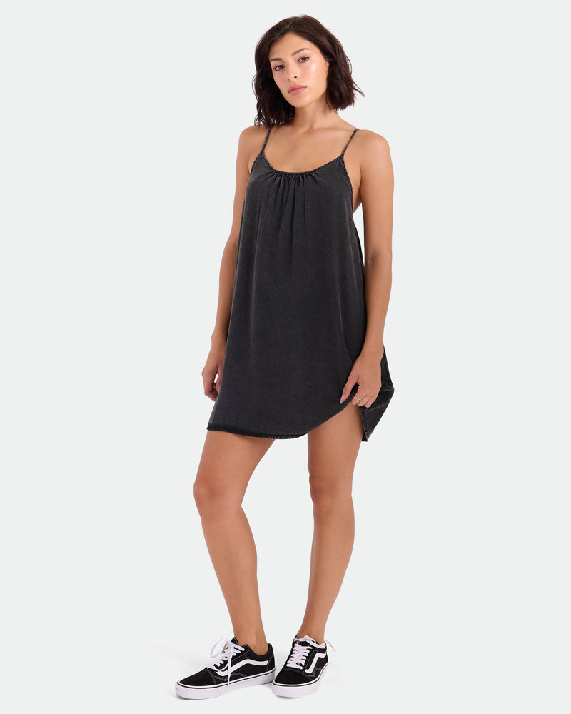 A woman wearing a loose, sleeveless Women's Baby Cakes Mini dress by Volcom with an adjustable tie-back and black Vans sneakers stands against a plain white background, looking at the camera with a neutral expression.
