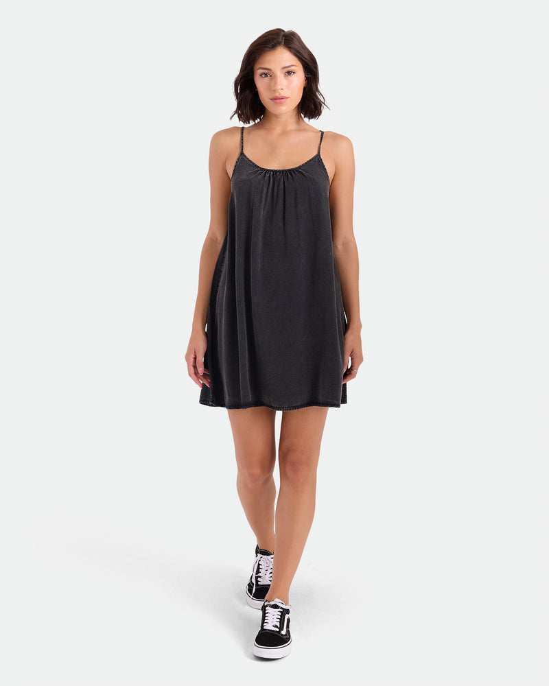 A woman with short brown hair wears the Volcom Women's Baby Cakes Mini, a vintage black woven day dress with thin straps and an adjustable tie-back, paired with black sneakers as she walks against a plain white background.