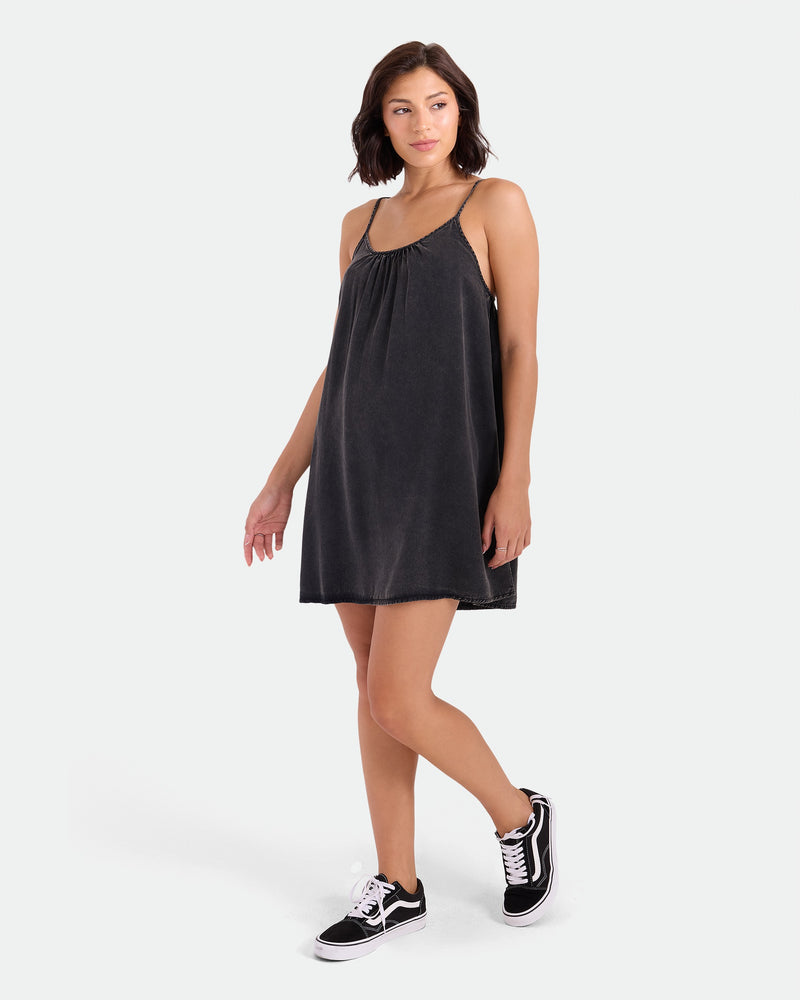 A woman with short brown hair models the Volcom Women's Baby Cakes Mini, a black woven dress with thin straps and an adjustable tie-back, paired with black Vans sneakers. She stands on a white background, gazing to the side with one leg forward.