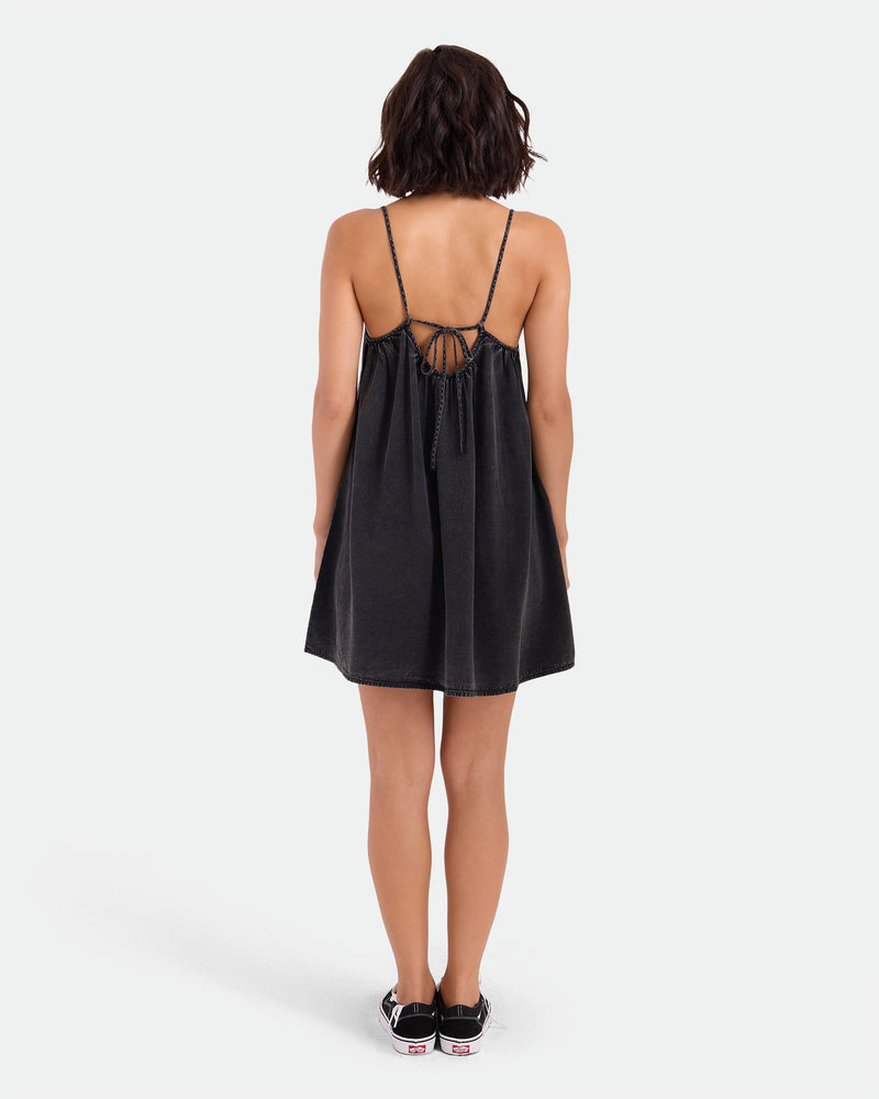 A woman with shoulder-length dark hair and black sneakers faces away, modeling the Volcom Women's Baby Cakes Mini—a Vintage Black woven day dress with adjustable tie-back straps and an open back—against a plain white background.