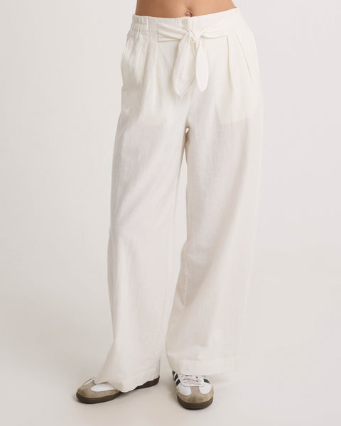 A person wears Volcom Women's Bow Chicka Pants—loose-fitting, high-waisted, cotton-linen wide legs with a tie-front waist—paired with white and gray sneakers. Photo cropped at the waist on a plain white background.